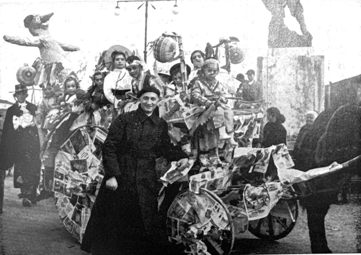 Don Livio Costagli, in tonaca, accanto a una vecchia lettiga addobbata con carta colorata e carica di bambini mascherati; fotografia in bianco e nero
